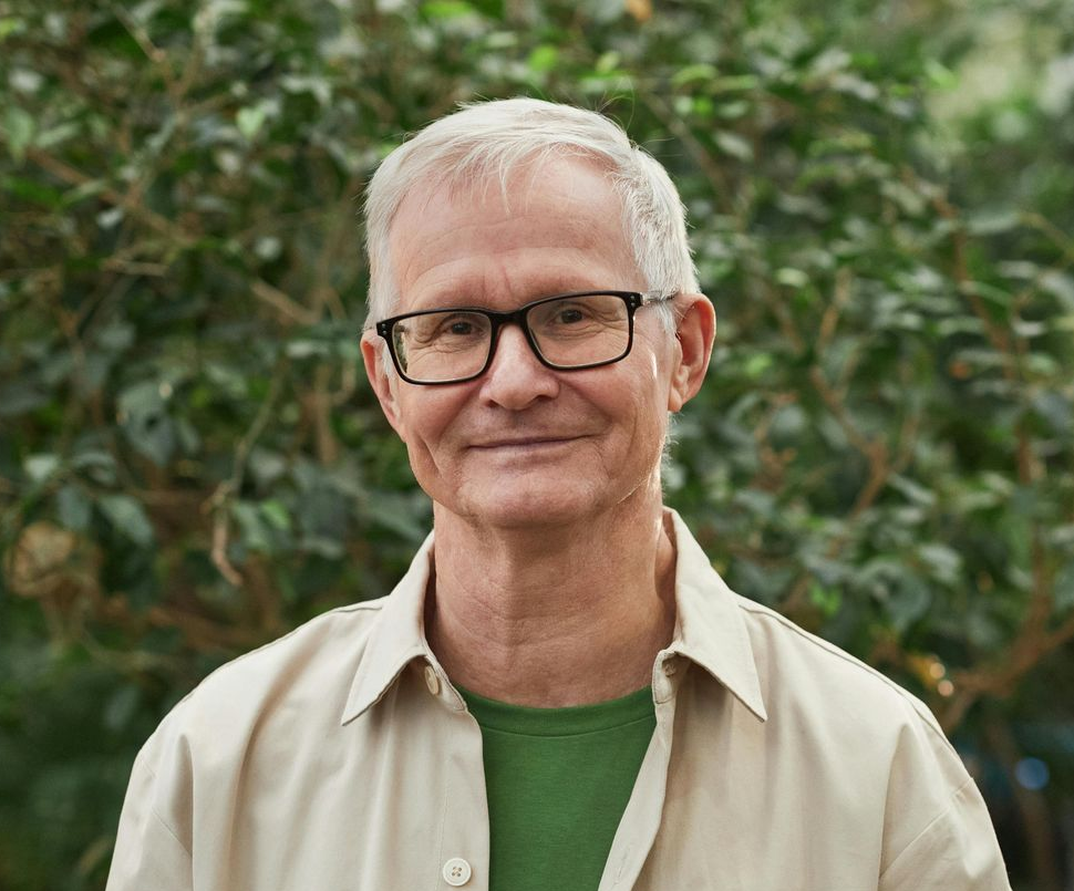 A man in a light blue shirt is standing in front of a window with his arms crossed.