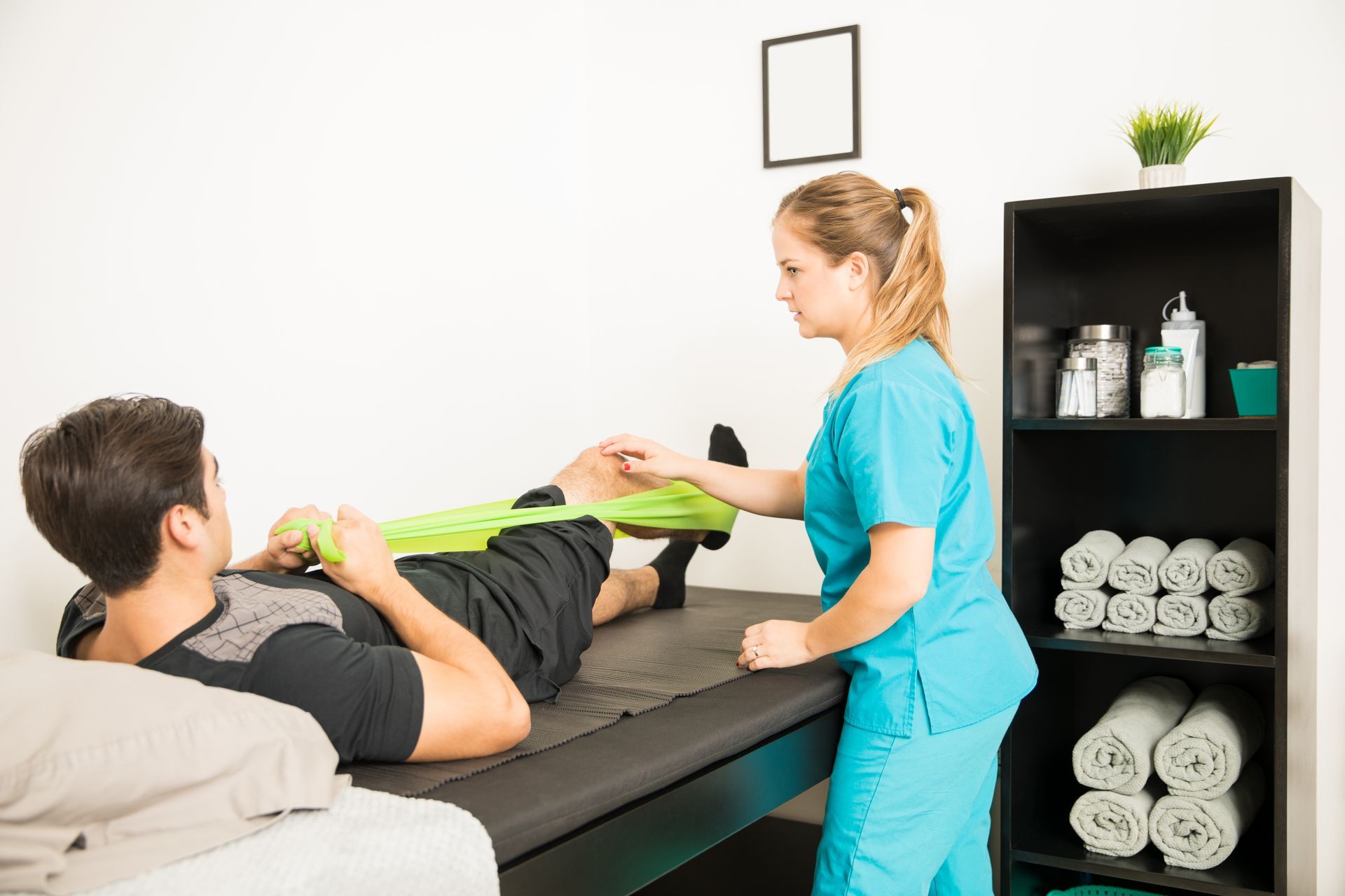 A therapist uses a resistance band to help a patient stretch his knee in a treatment room.