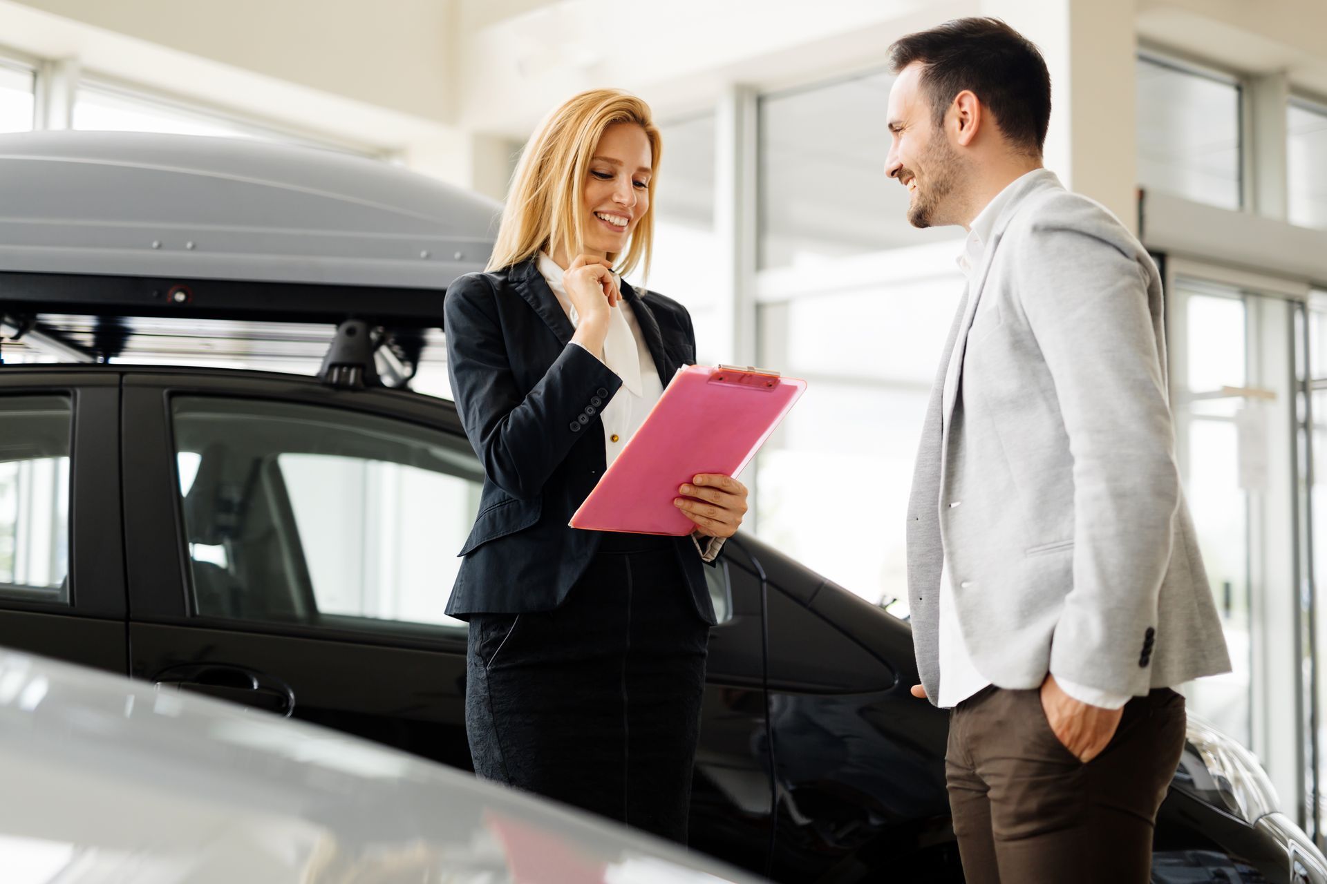 Woman in a suit showing a clipboard to a man, standing next to a car in a dealership.