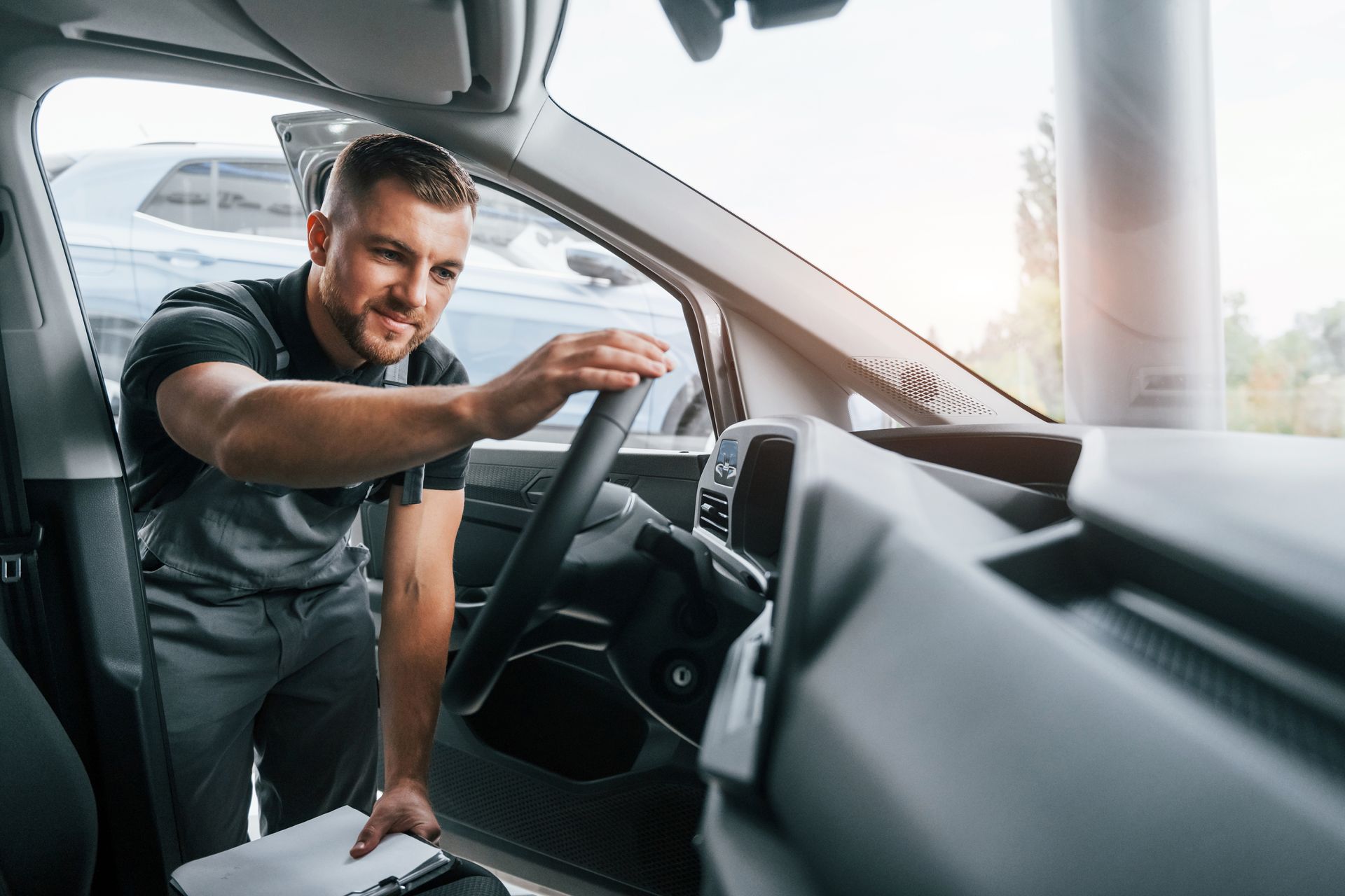 Mechanic in grey overalls inspecting car dashboard, holding clipboard.