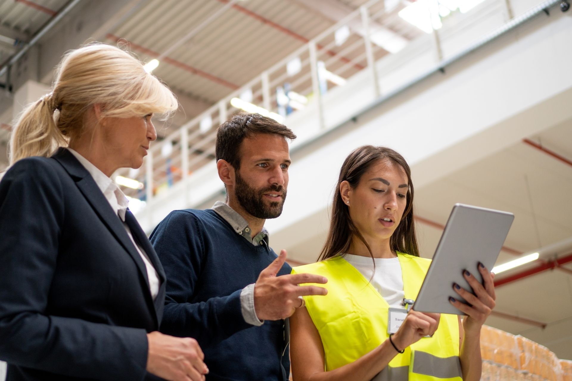 Three people in a warehouse looking at a tablet. One wears a yellow vest, others are in business attire.