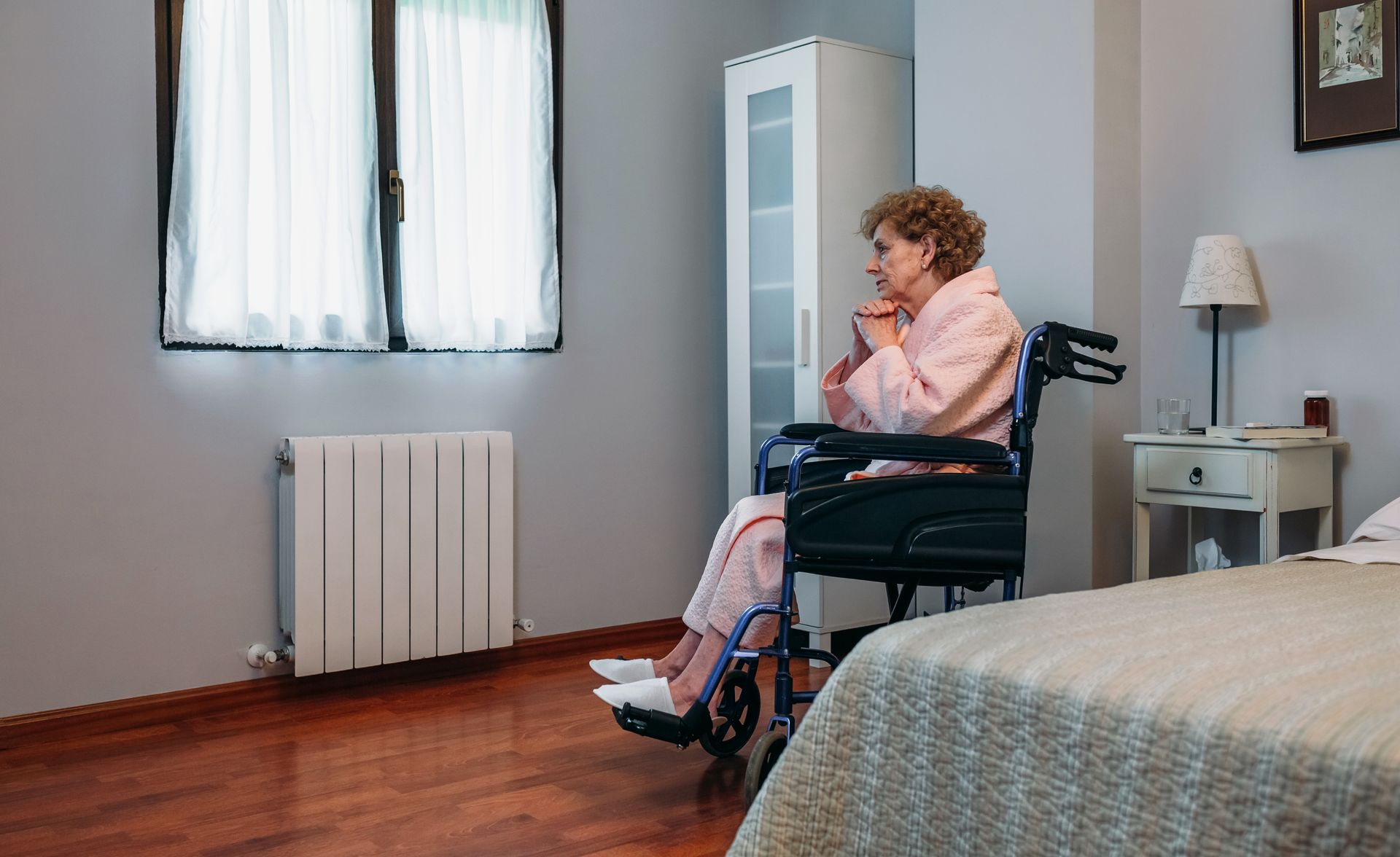 Woman in wheelchair sits by window in bedroom, looking thoughtful.