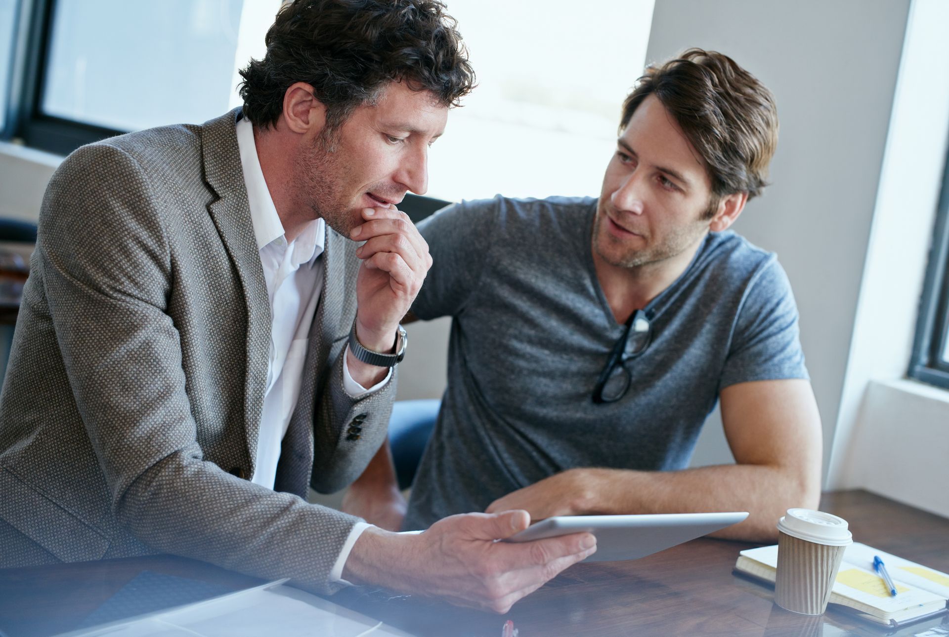 Two men looking at tablet, one pointing, discussing ideas. Coffee cup and notepad nearby.