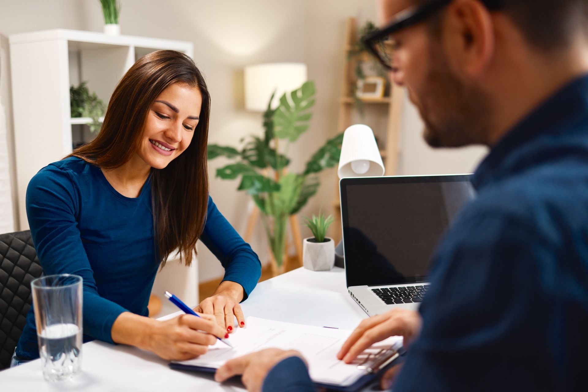 Woman signing papers at a desk with another person, laptop, and water glass present; indoor setting.