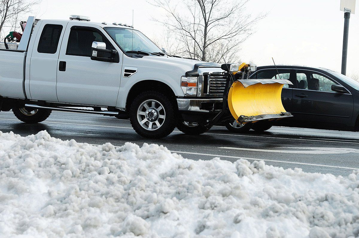 White pickup truck with snowplow pushing snow alongside a parked car.