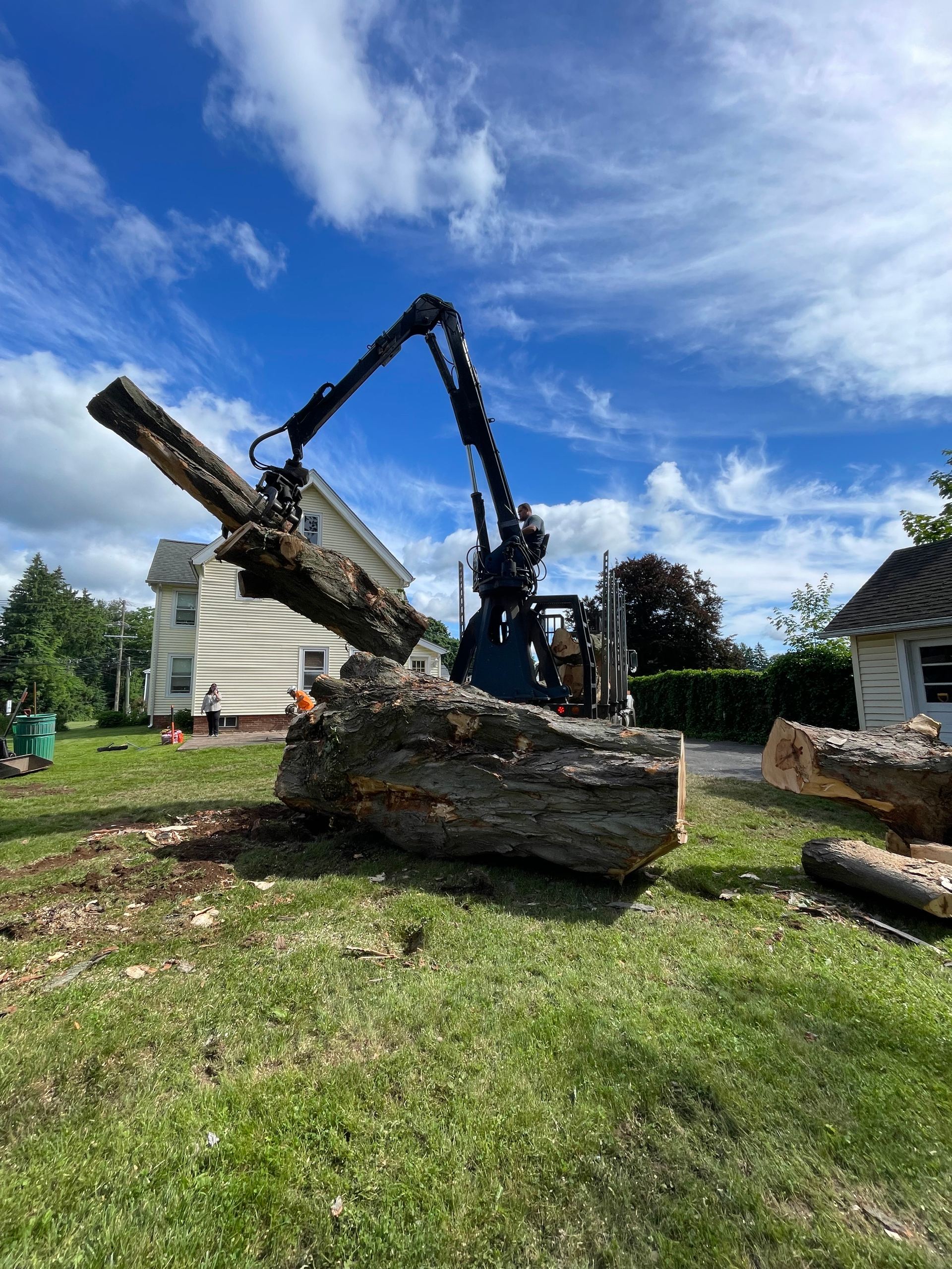 A large tree log being lifted by a blue forestry machine on a grassy lawn. A house is in the background under a blue sky.