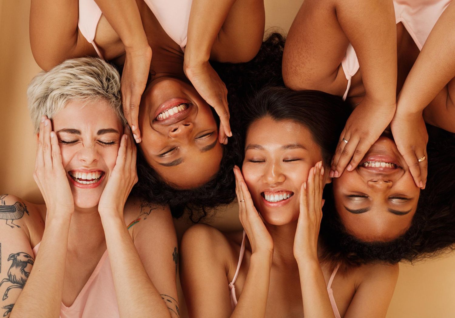 Four diverse women smiling, hands on their faces. They are lying down, wearing pink tops, against a neutral background.