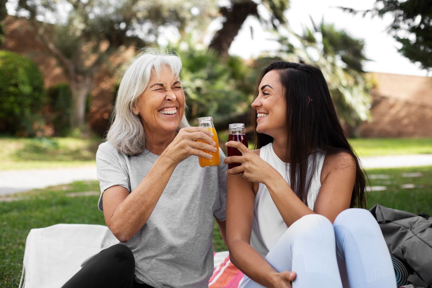 Two smiling women toasting with juice bottles outdoors in a park.