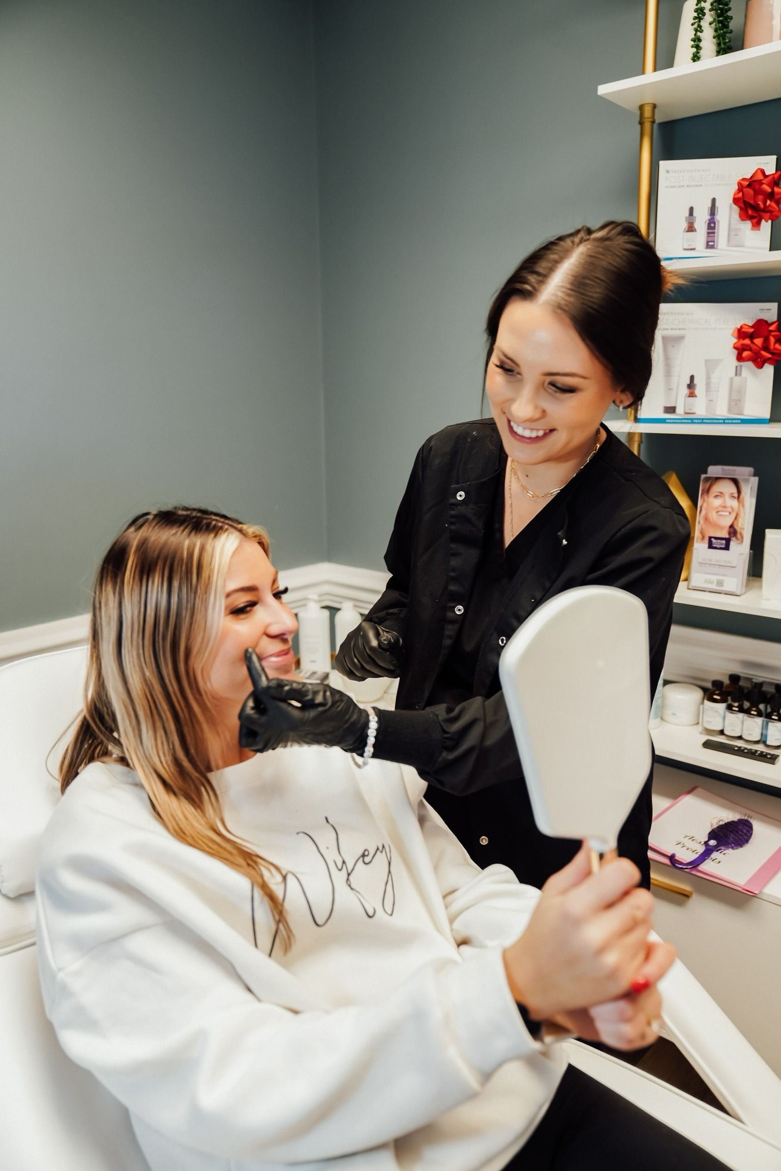 Woman receiving beauty treatment, looking at a mirror, smiling. Aesthetician in black scrubs smiles too, in a beauty clinic.