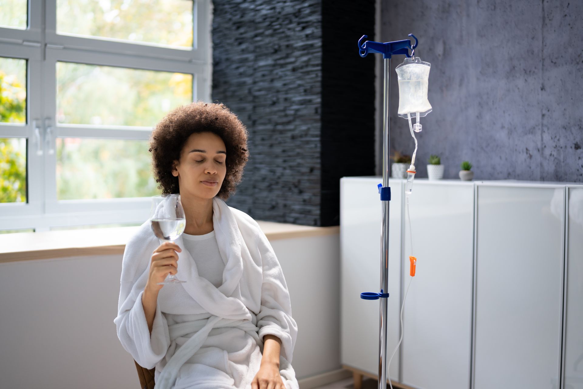 Woman in robe receiving IV fluids, sitting by a window, with a white cabinet in the background.