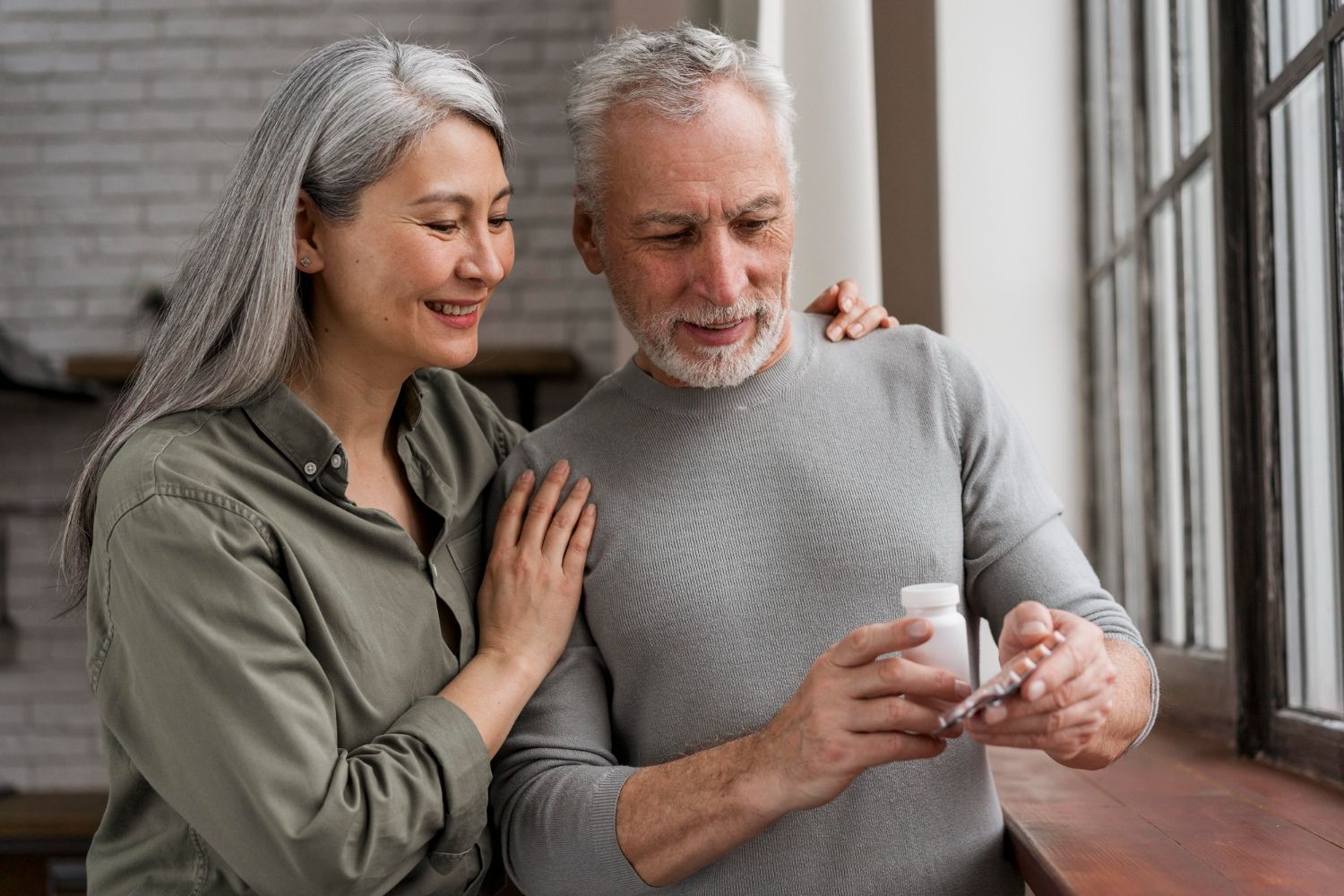 Elderly couple looking at pills near a window; woman has arm around man, both with gray hair.