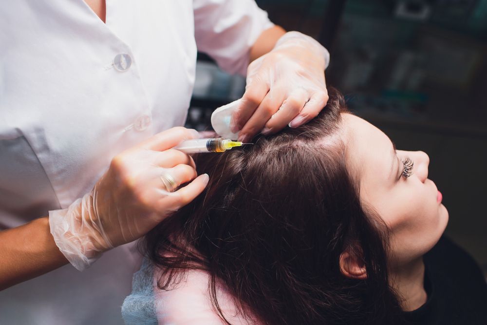 A woman receives a scalp injection; a medical professional in gloves holds a syringe near her hair.