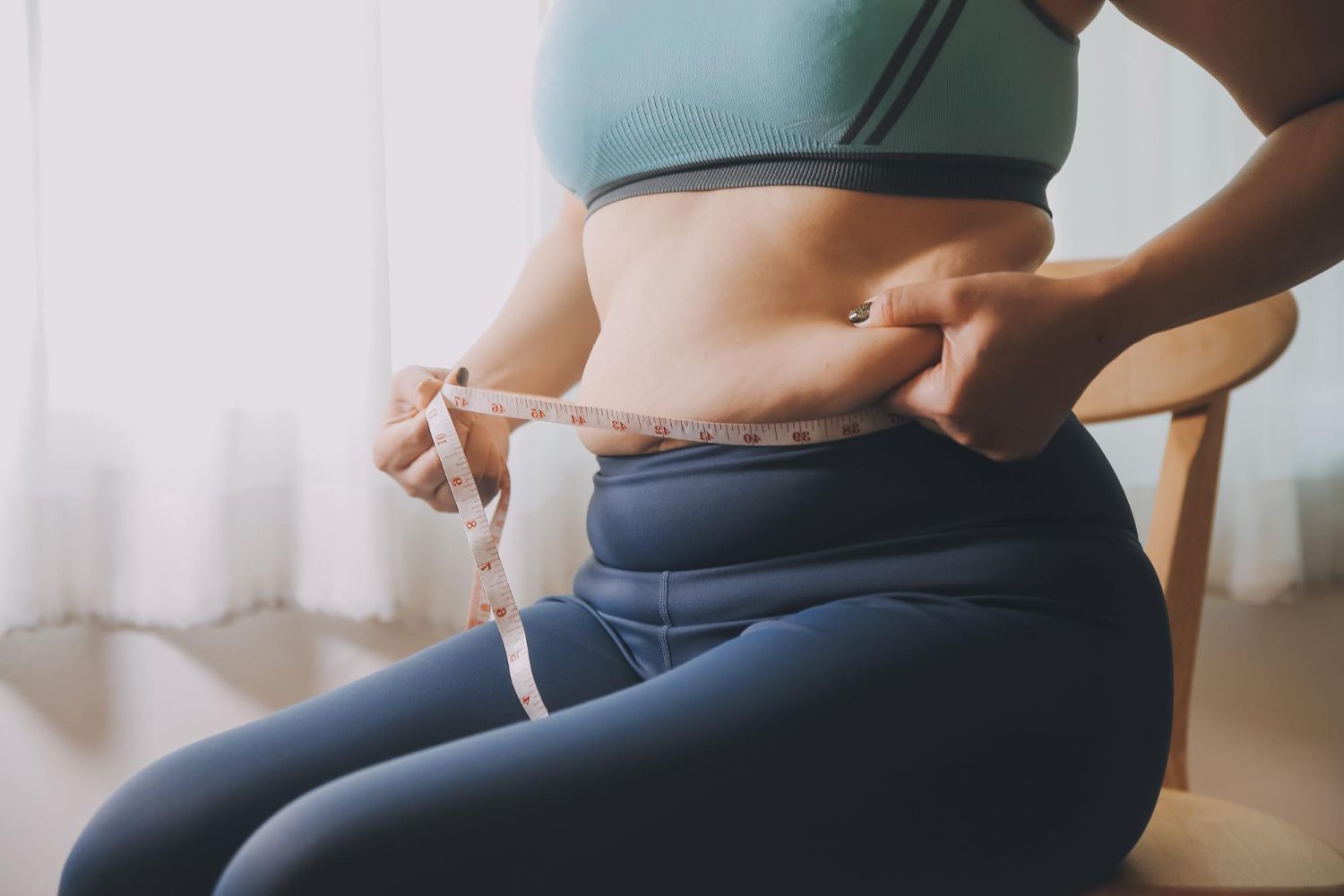 Woman in workout clothes measuring her waist with a tape measure while sitting on a chair.