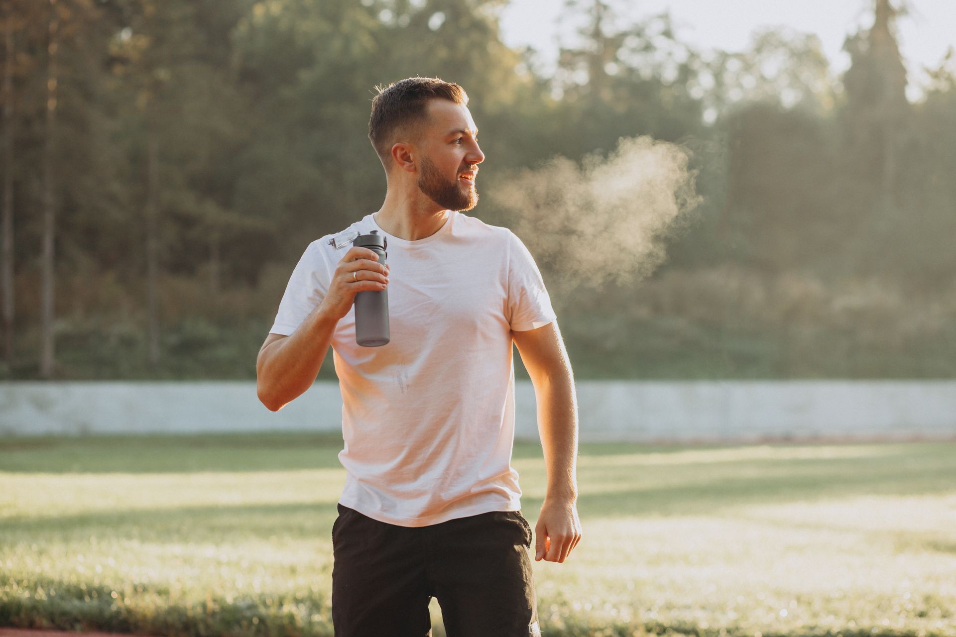 Man in white shirt and black shorts exhaling mist, holding water bottle on a track.