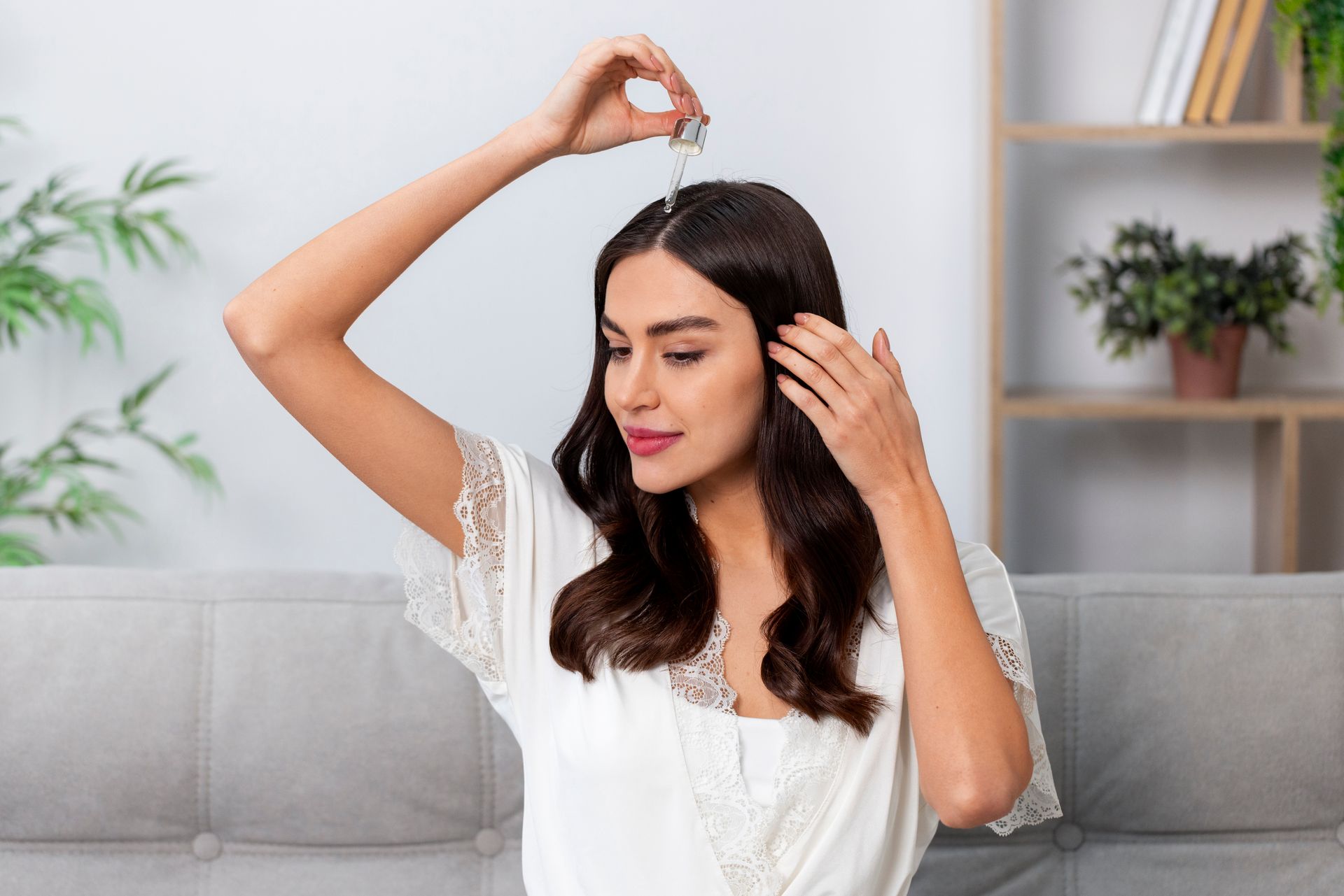 A woman is applying a serum to her hair while sitting on a couch.