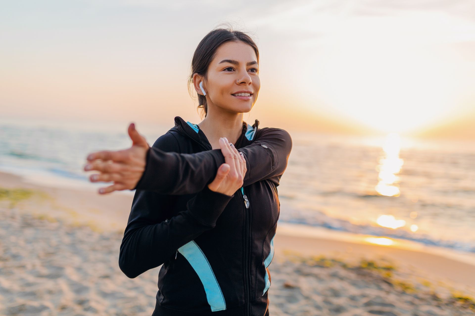 Woman stretches arm at beach with ocean and sunrise in background.