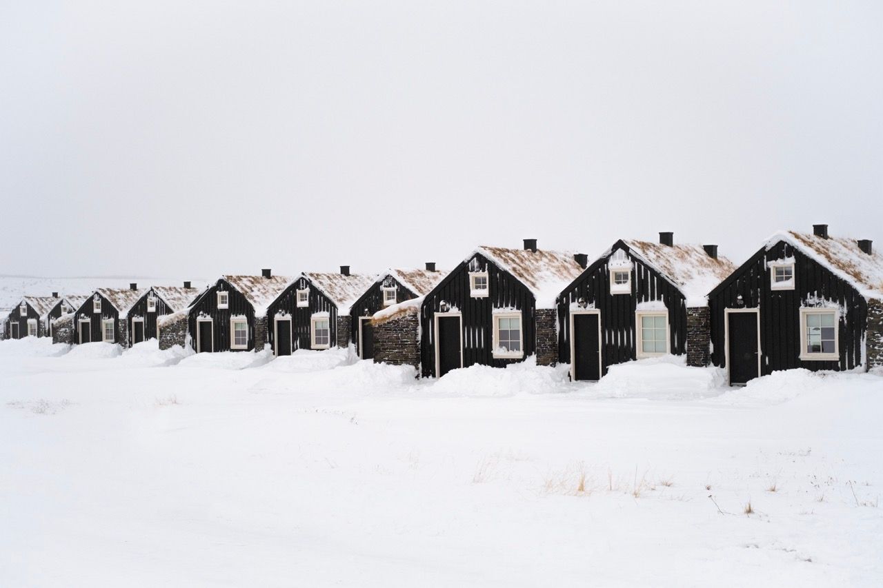 Three small houses are sitting in the middle of a field with mountains in the background.