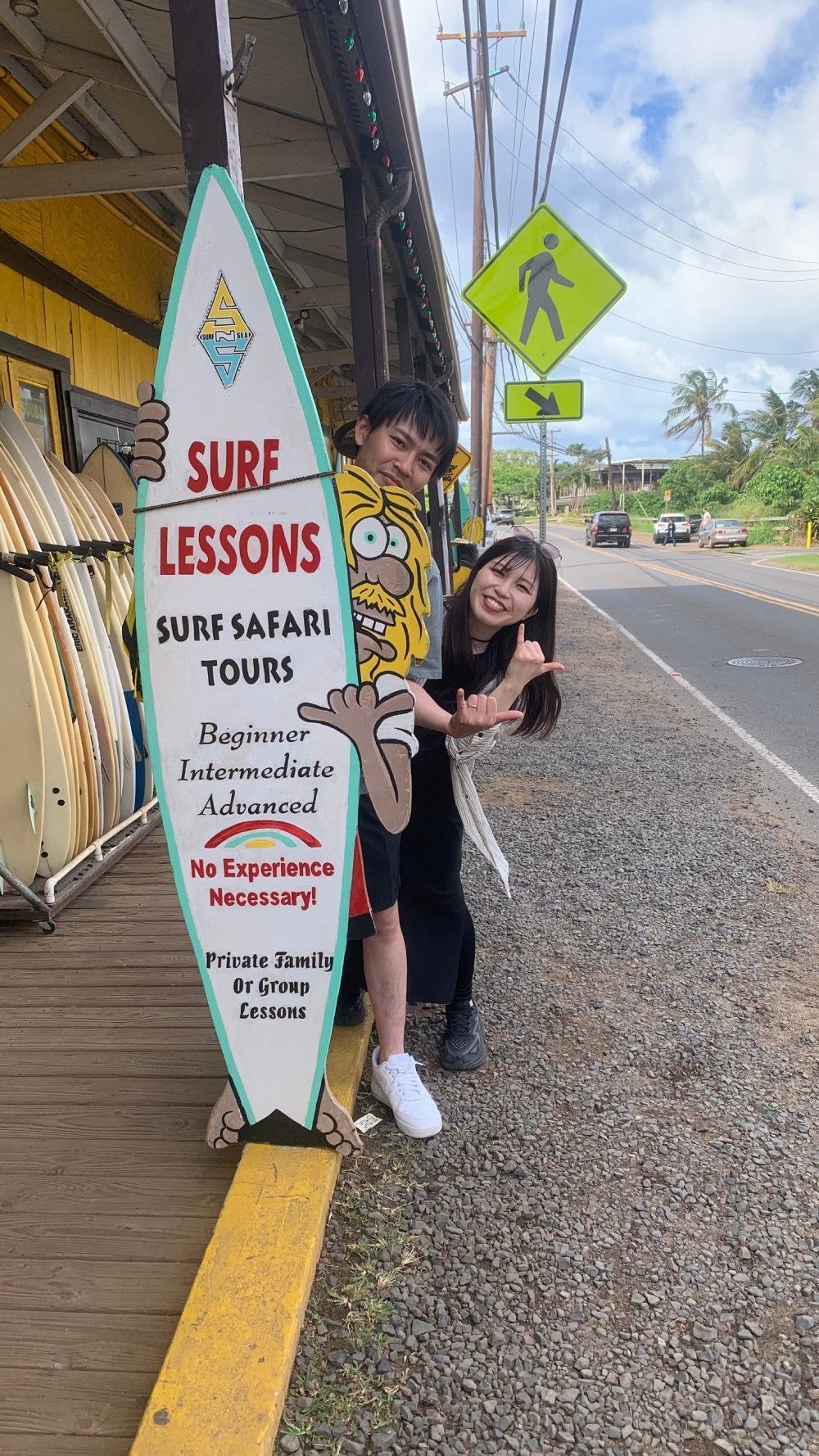 Two people holding a surfboard sign advertising surf lessons on a sidewalk near a road.