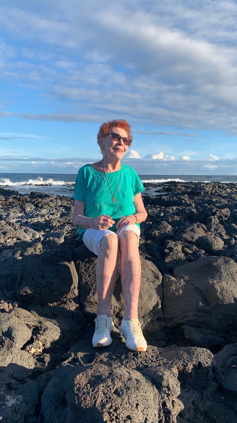 An elder woman is sitting on a rock near the ocean