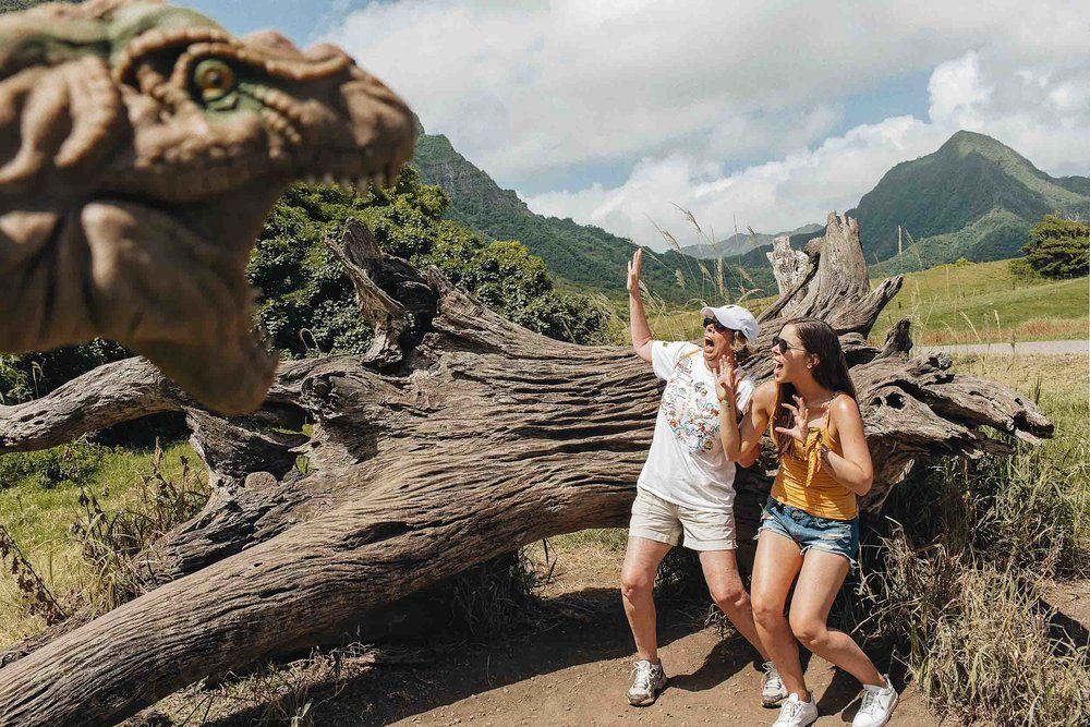 Two women are sitting on a log in front of a dinosaur statue.