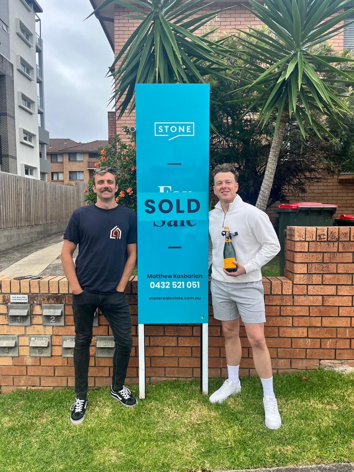 Man with glasses hugs woman from behind; both smile outdoors.