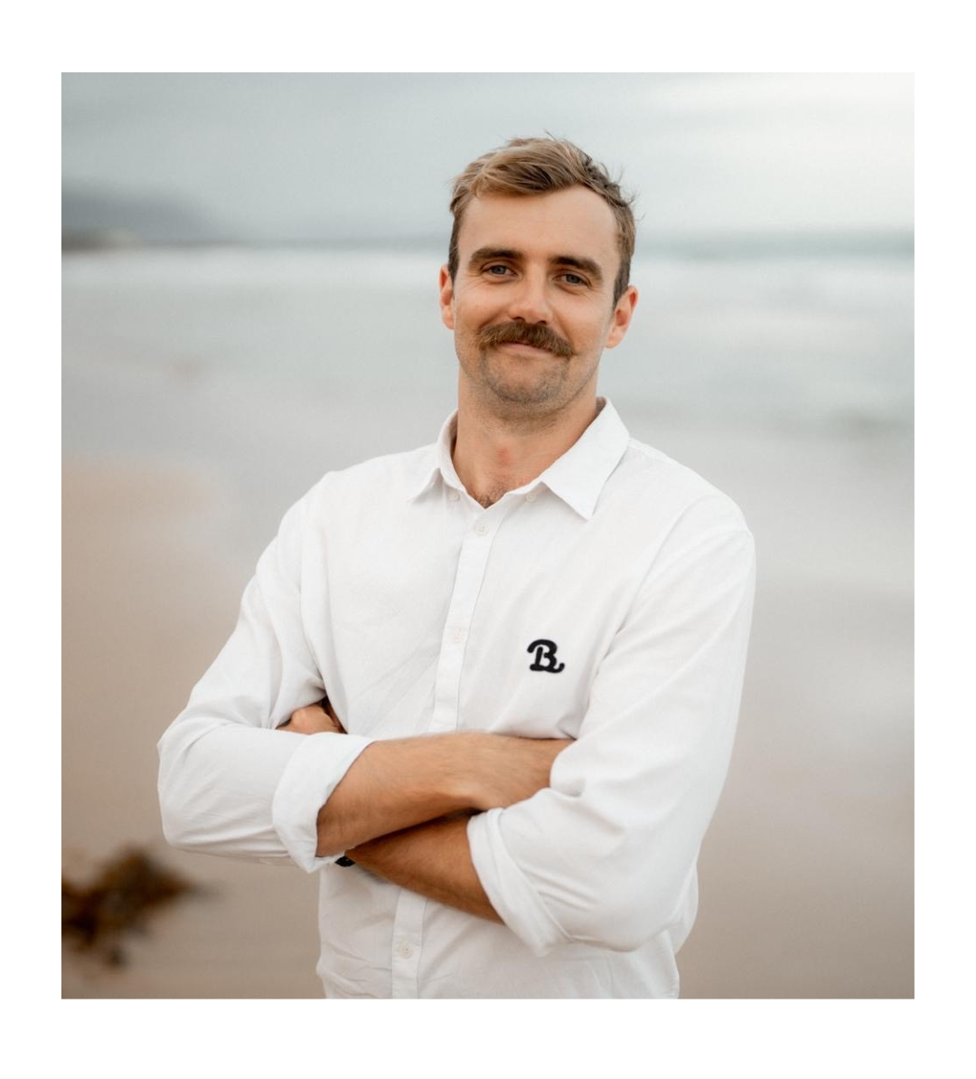 Man with mustache, arms crossed, standing on a beach. Wearing a white shirt with logo.