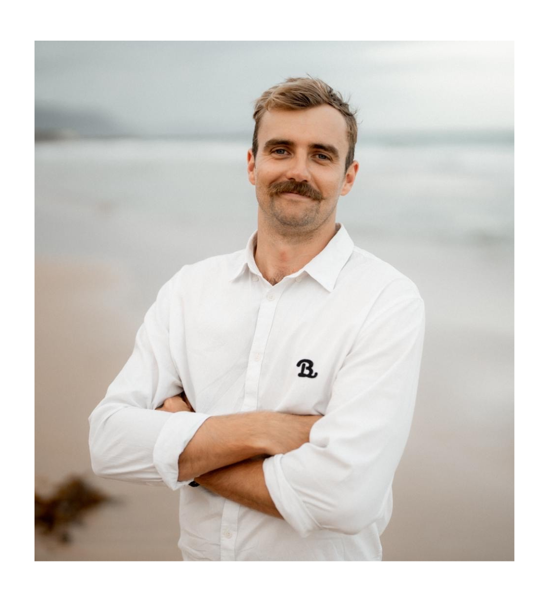 Man with mustache in a white shirt, arms crossed, standing on a beach with a blurred ocean background.