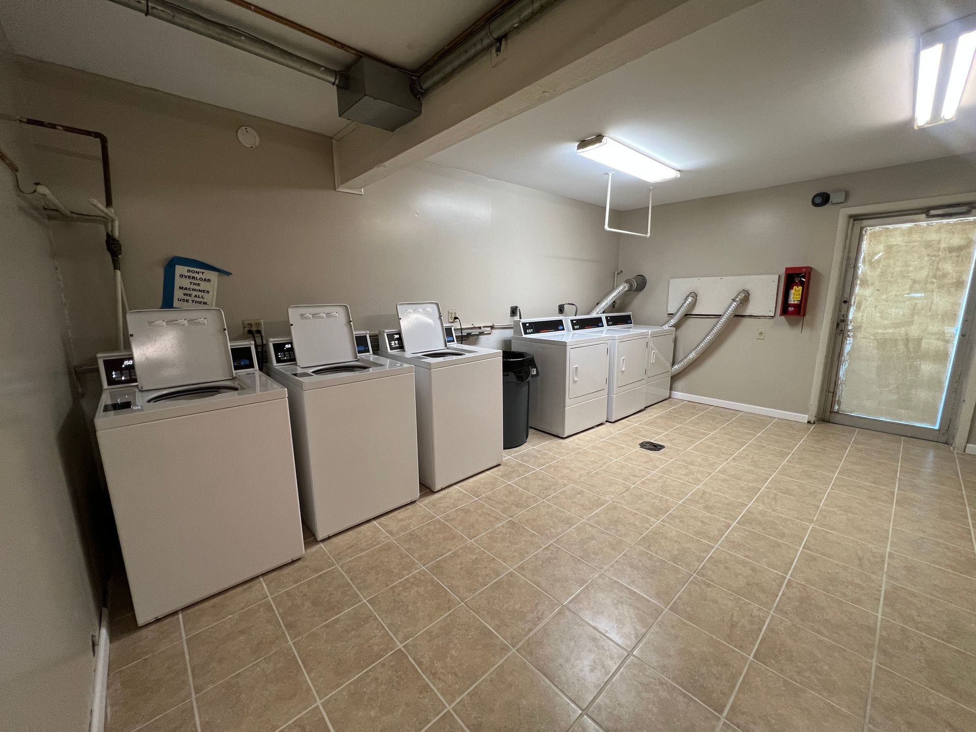 Laundry room with multiple washing machines and dryers. Tiled floor, neutral walls, and a door to the outside.