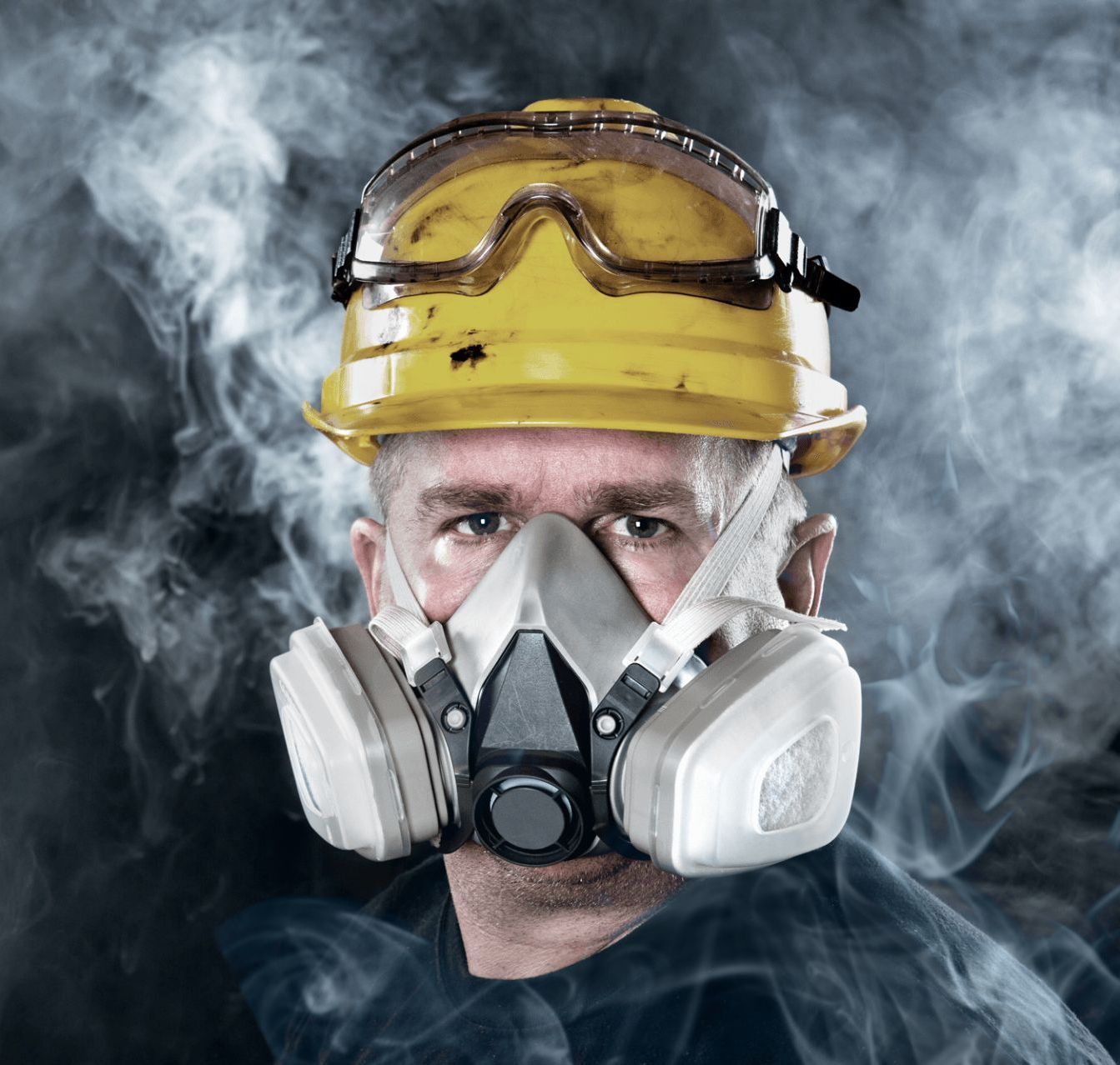 A man wearing a hard hat and gas mask is surrounded by smoke.