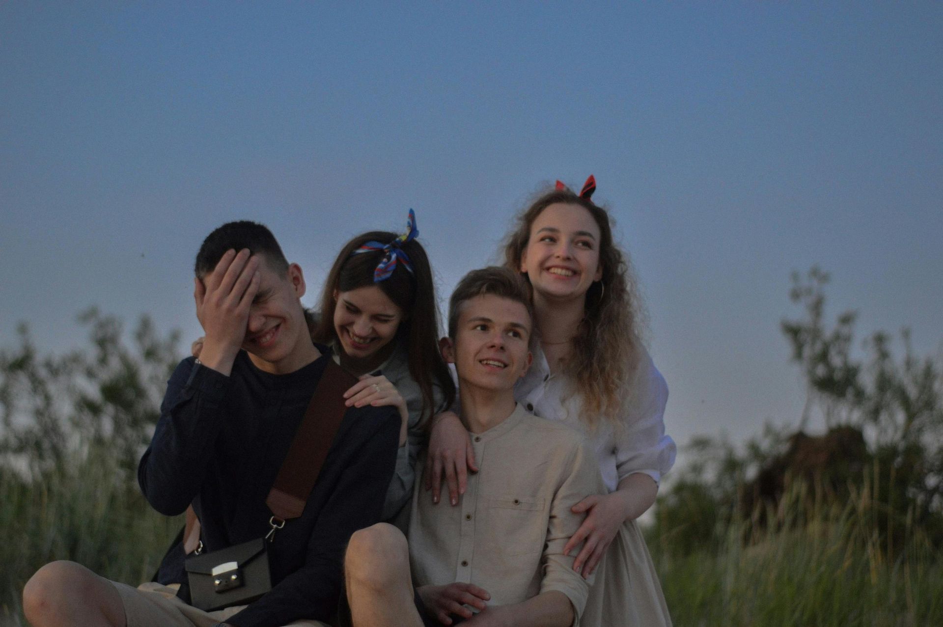 Four friends sitting in grass, smiling and hugging at dusk under a clear sky