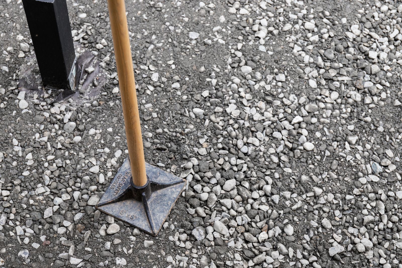 A hand tamper with a wooden handle is resting on a gravel surface next to a black post.