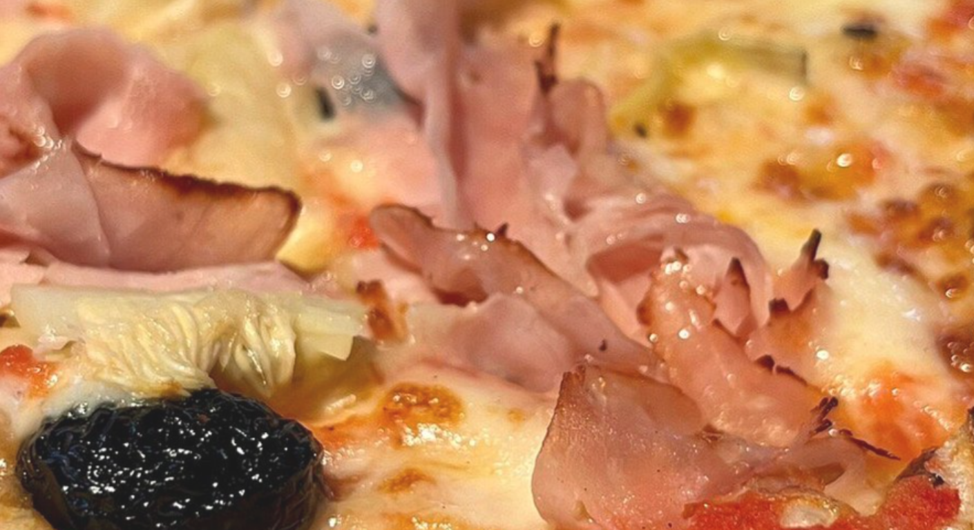 Primo piano di una pizza condita con fette di prosciutto, formaggio fuso e un'oliva nera.