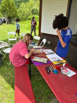 Children crafting at a red picnic table outdoors beside a house, with art supplies and folding chairs nearby.