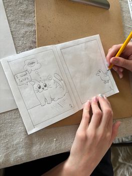 Hands on an open workbook, sketching with a pencil beside a brown folder.