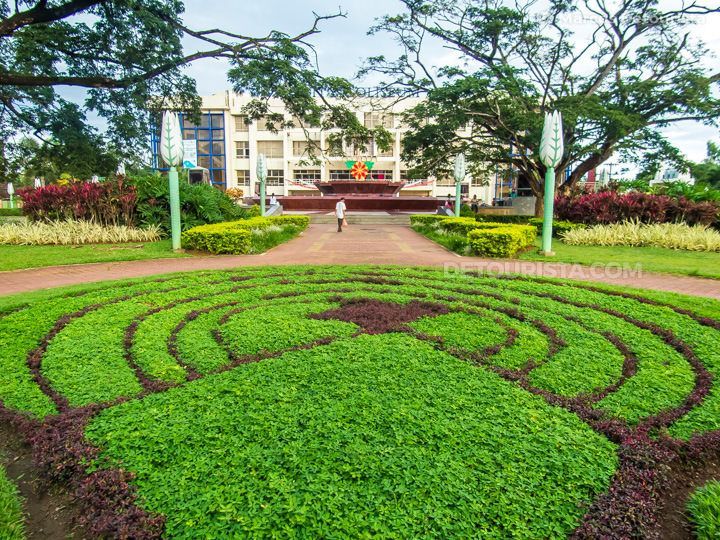 Park with spiral plant beds leading to a building with a flag; person walking.