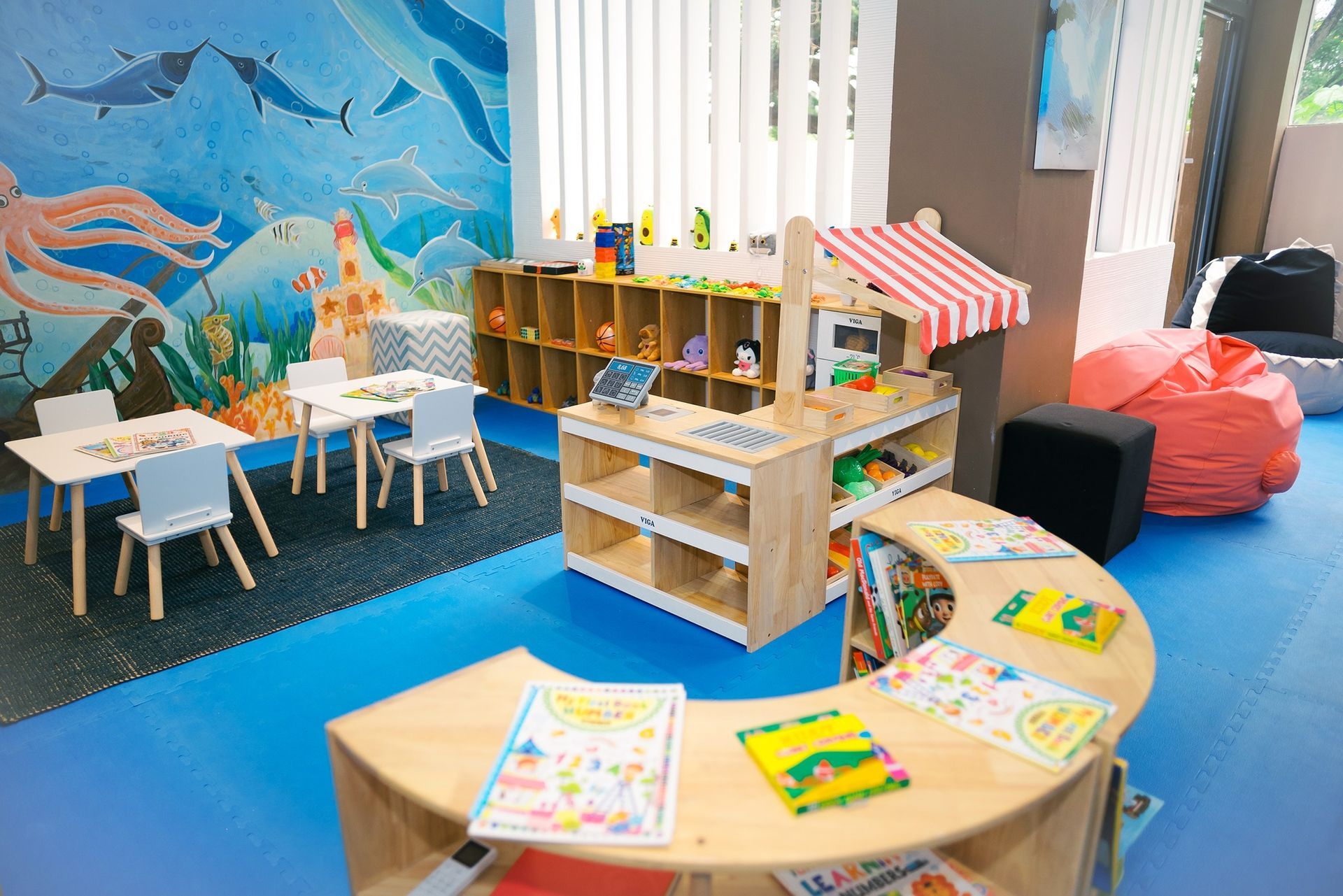 A children's playroom with blue flooring and ocean-themed wall art. Play tables, shelves of toys, and a reading nook are present.