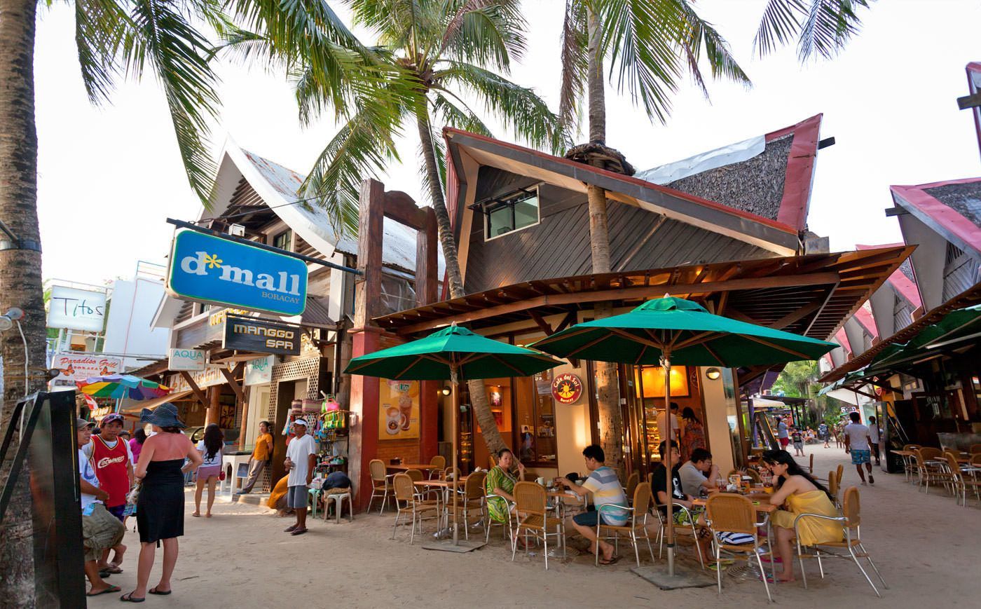 People are sitting at tables outside D'Mall with umbrellas.
