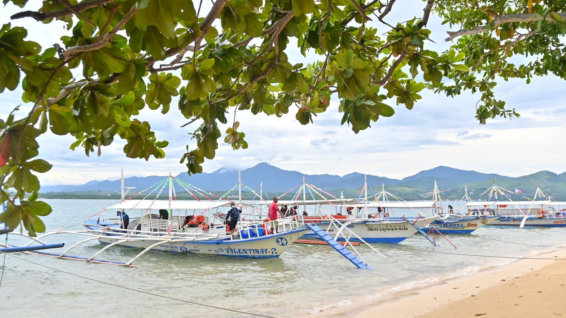 A group of boats are docked on a beach with mountains in Honda Bay