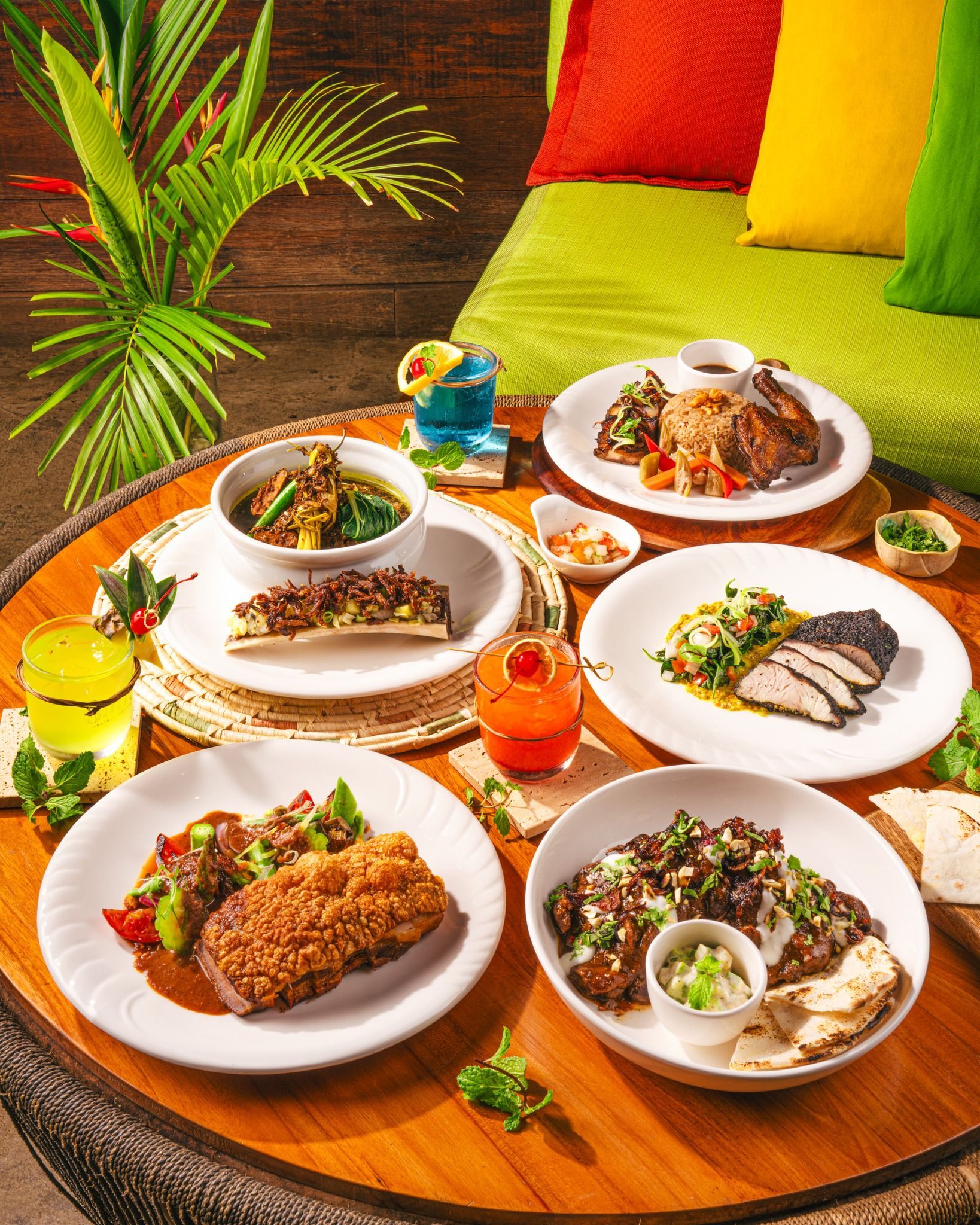 Platter of various dishes and cocktails on a table, with tropical foliage in the background.
