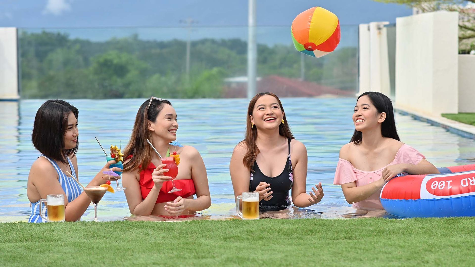 Four people in a pool, smiling and holding drinks. A beach ball floats above them.