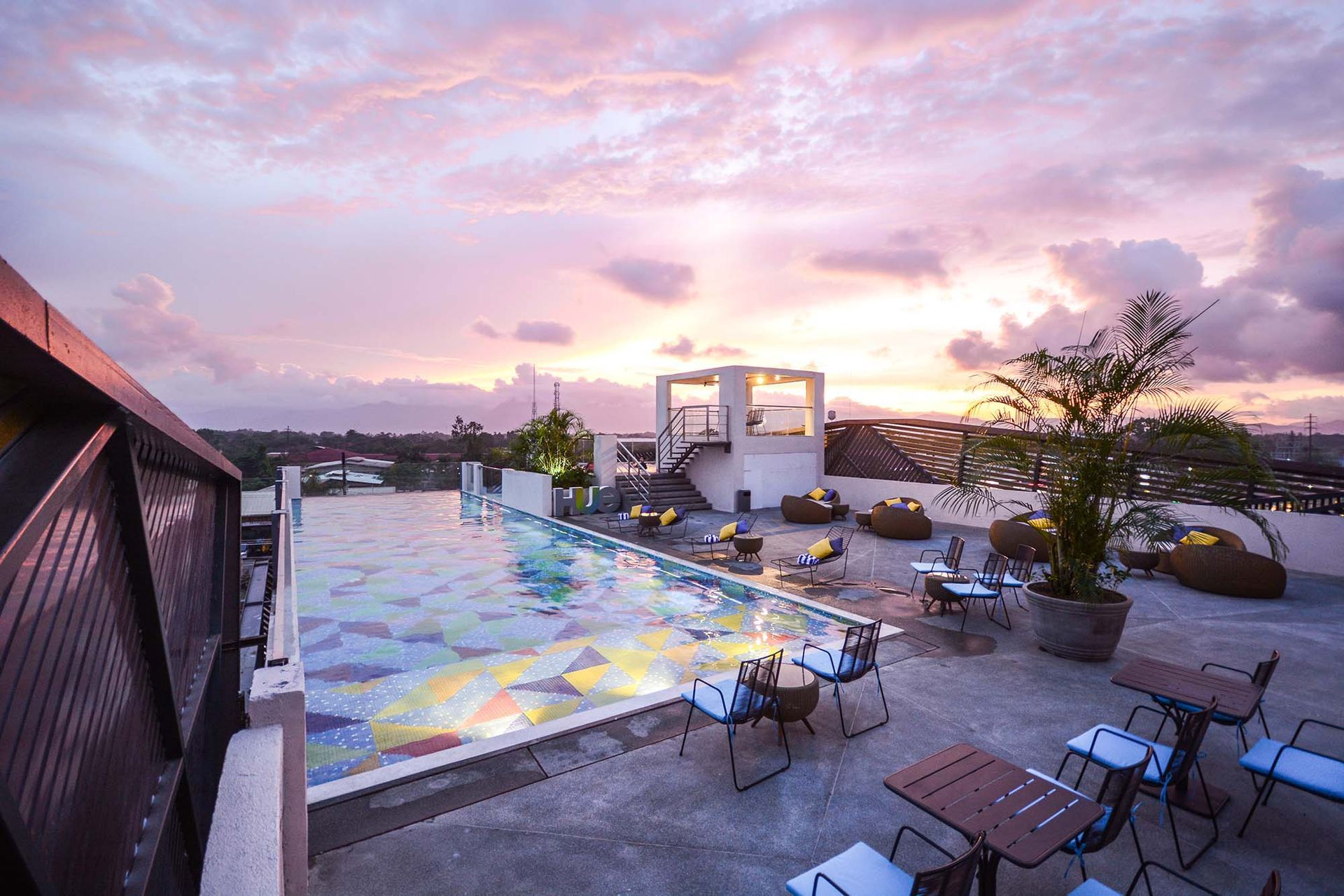 Rooftop swimming pool with tables and chairs around it at sunset.