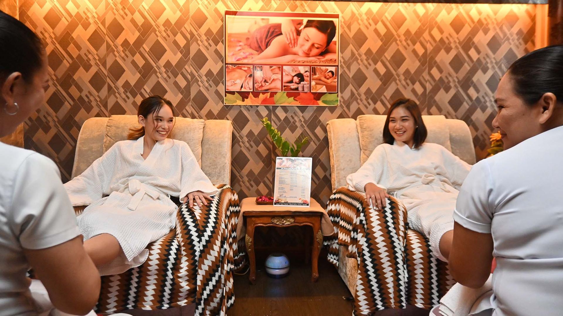 Two people receive foot massages in a spa, smiling. They sit in cushioned chairs, covered by blankets. Two therapists work on their feet.