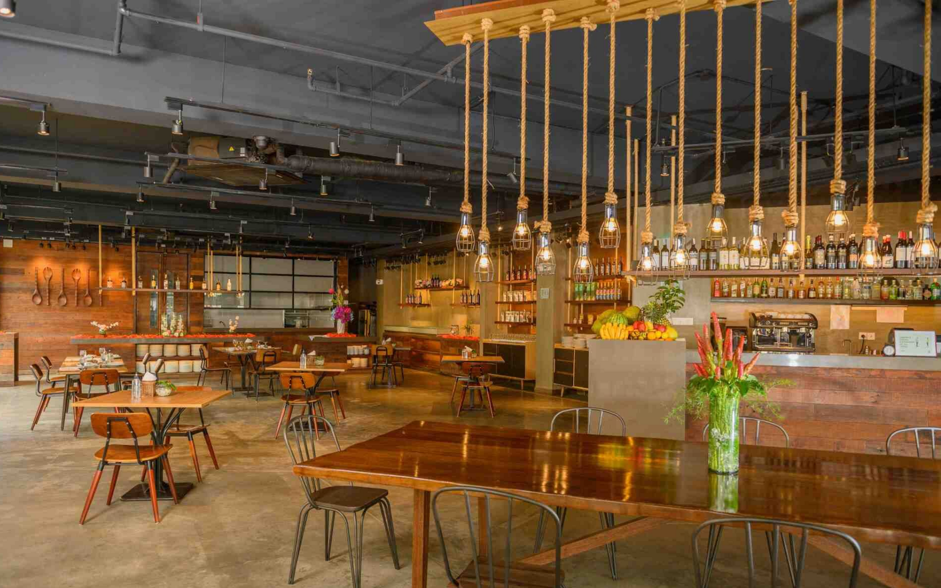 A spacious restaurant dining area featuring wooden tables, metal chairs, and low-hanging Edison bulb light fixtures.