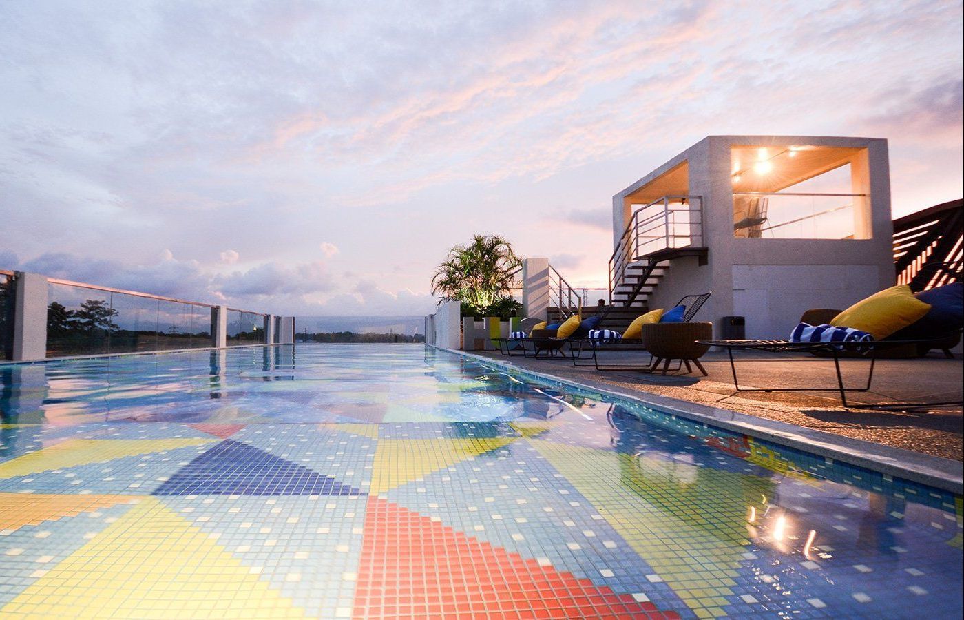 Rooftop pool with colorful mosaic tiles, chairs, and a white building under a sunset sky.