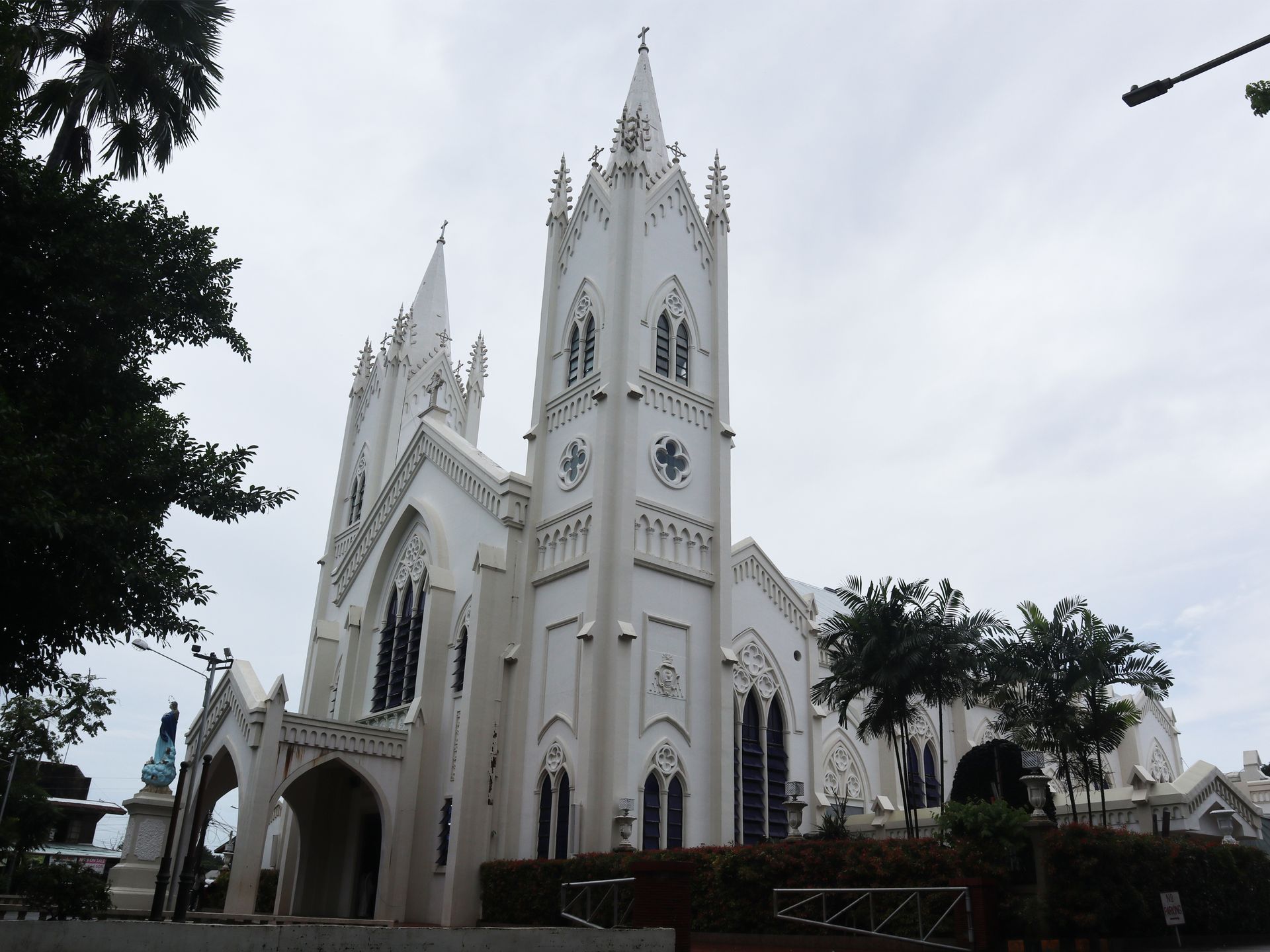 A white, Neo-Gothic Catholic church with twin spires stands against a cloudy sky, surrounded by trees and a stone wall.