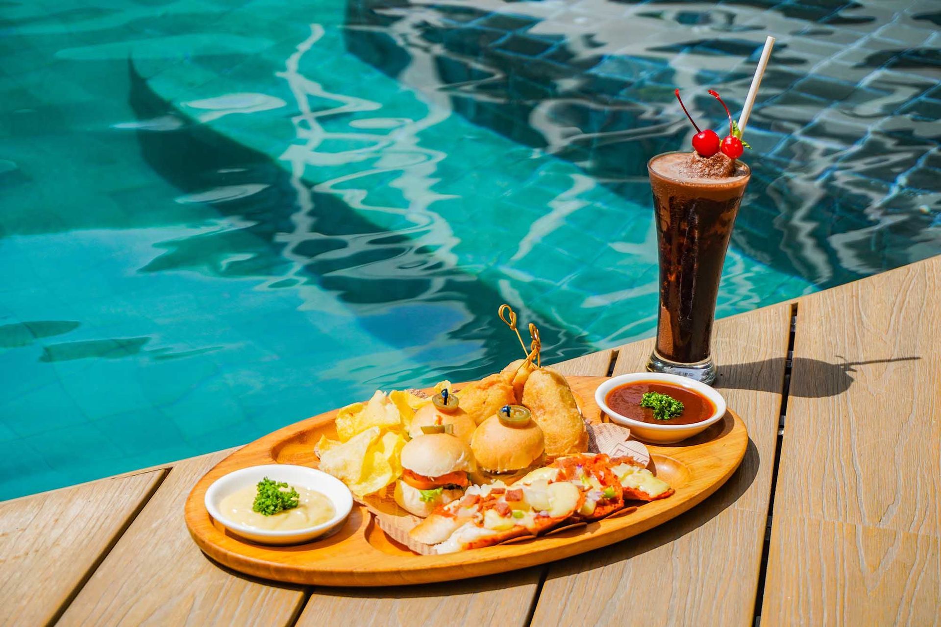 A wooden tray filled with food and a drink next to a swimming pool.