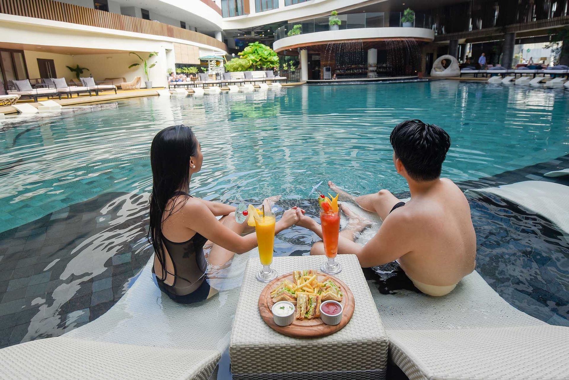 Couple relaxing in pool, drinks and snacks on a small table.