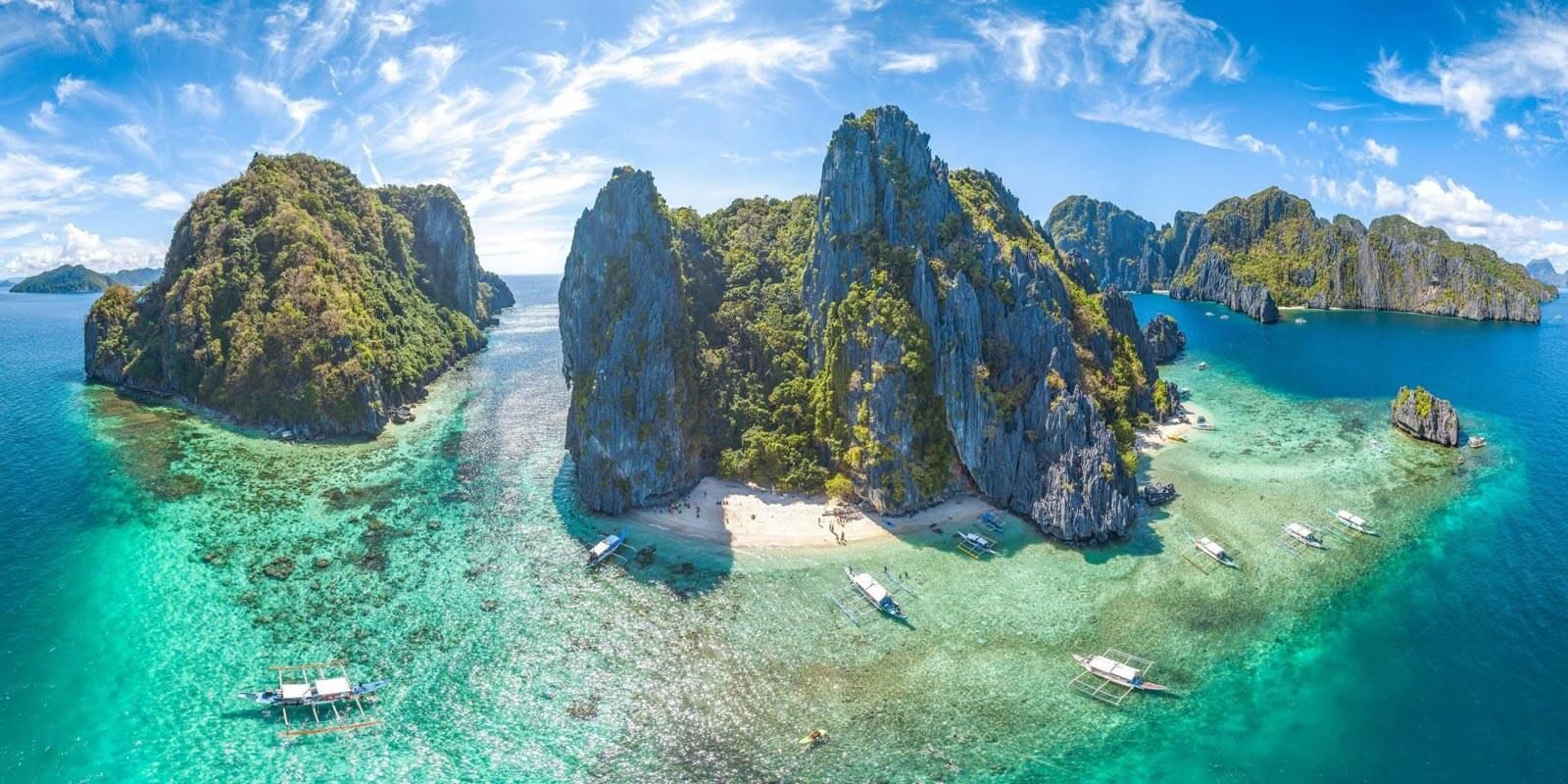 Island-filled ocean landscape with boats, turquoise water, and dramatic rock formations under a blue sky.