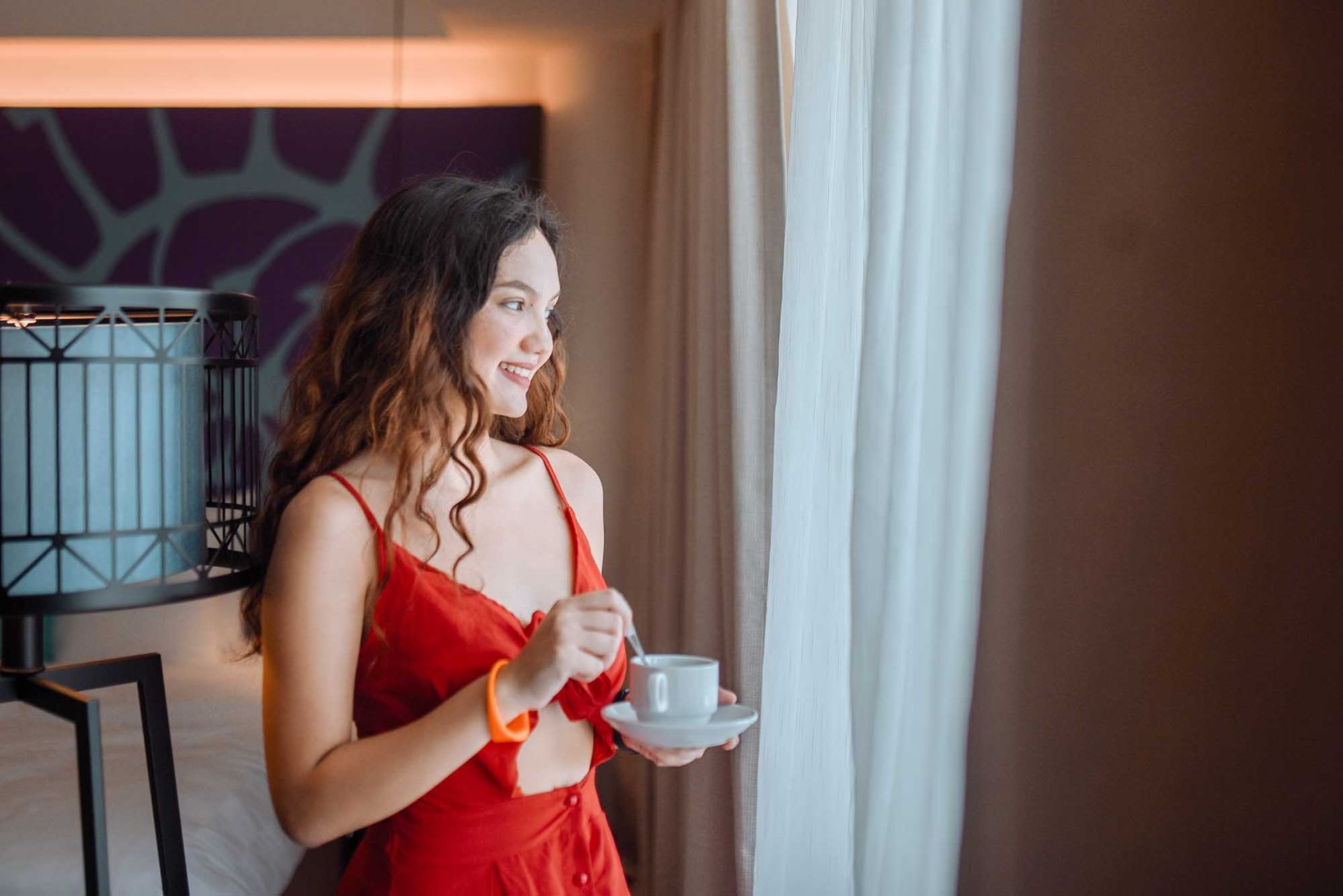 A woman in a red dress is holding a cup of coffee and looking out of a window.
