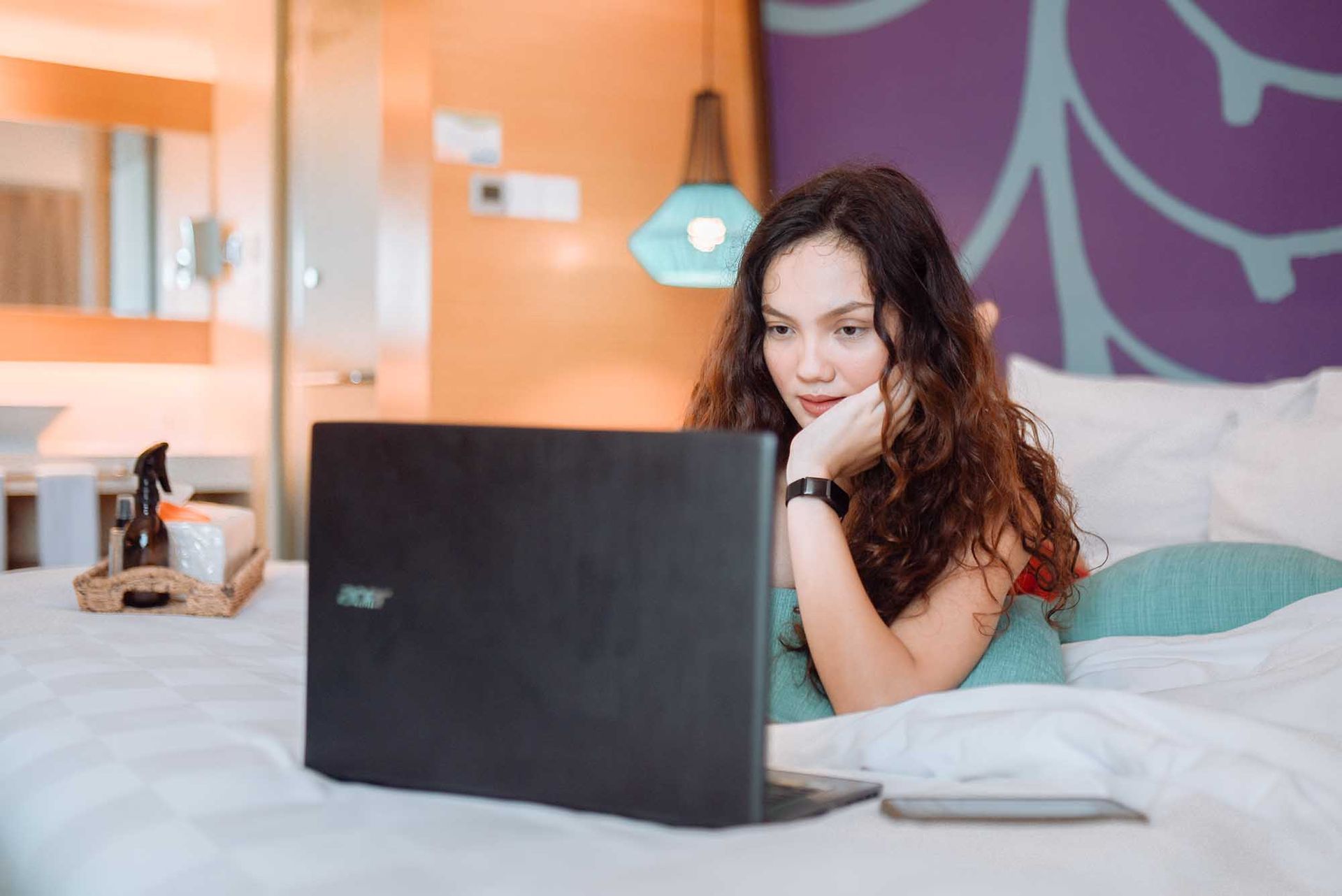Woman using a laptop while lying on a bed in a hotel room, looking focused.
